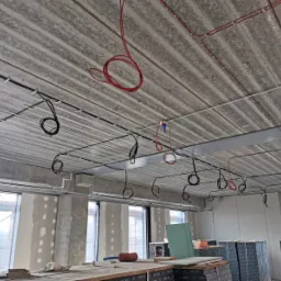 Surowy strop z widocznymi przewodami elektrycznymi w kolorze czarnym i czerwonym, zamocowanymi do sufitu za pomocą uchwytów, w tle okna i fragment rusztowania.