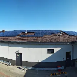 Instalacja paneli na dachu budynku przemysłowego z brązową dachówką, widok z góry, błękitne niebo w tle.