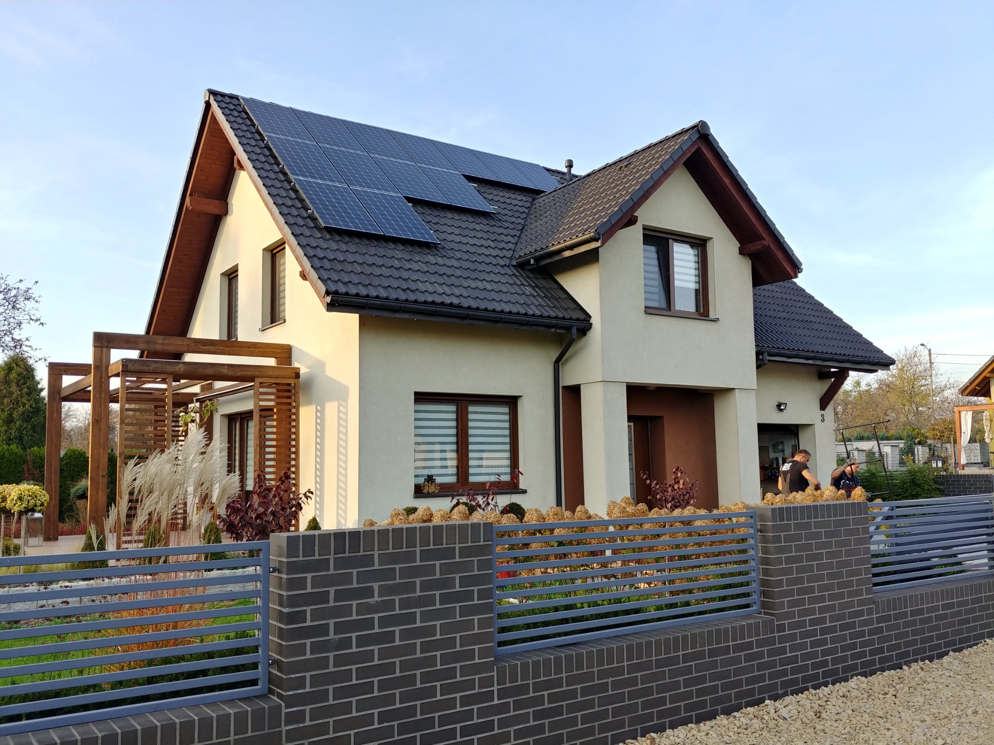 Nowoczesny dom jednorodzinny z panelami fotowoltaicznymi na dachu, widok zza grafitowego ogrodzenia z cegły klinkierowej.