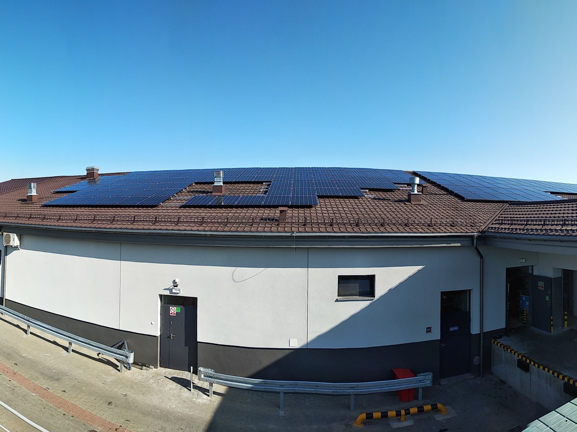 Instalacja paneli na dachu budynku przemysłowego z brązową dachówką, widok z góry, błękitne niebo w tle.