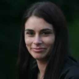 Portret uśmiechniętej kobiety z ciemnymi włosami, ubrana w ciemną marynarkę, na rozmytym zielonym tle.