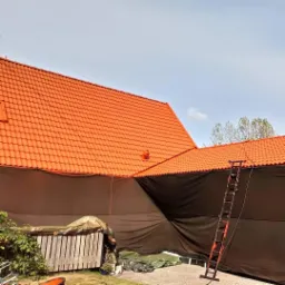 Pomarańczowy dach domu, częściowo zakryty czarną folią ochronną, z przystawioną drabiną, widok z dołu.