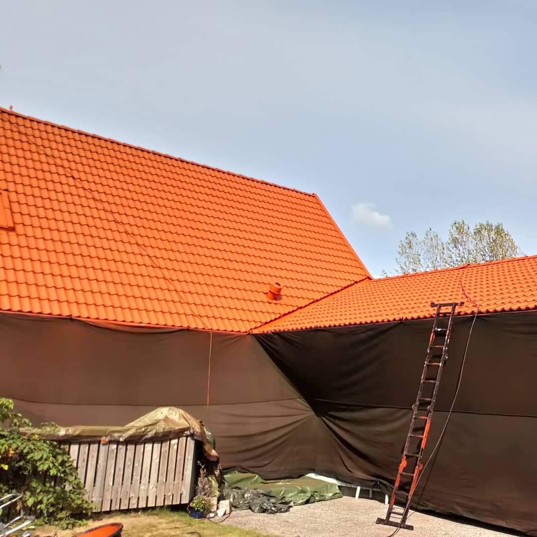 Pomarańczowy dach domu, częściowo zakryty czarną folią ochronną, z przystawioną drabiną, widok z dołu.