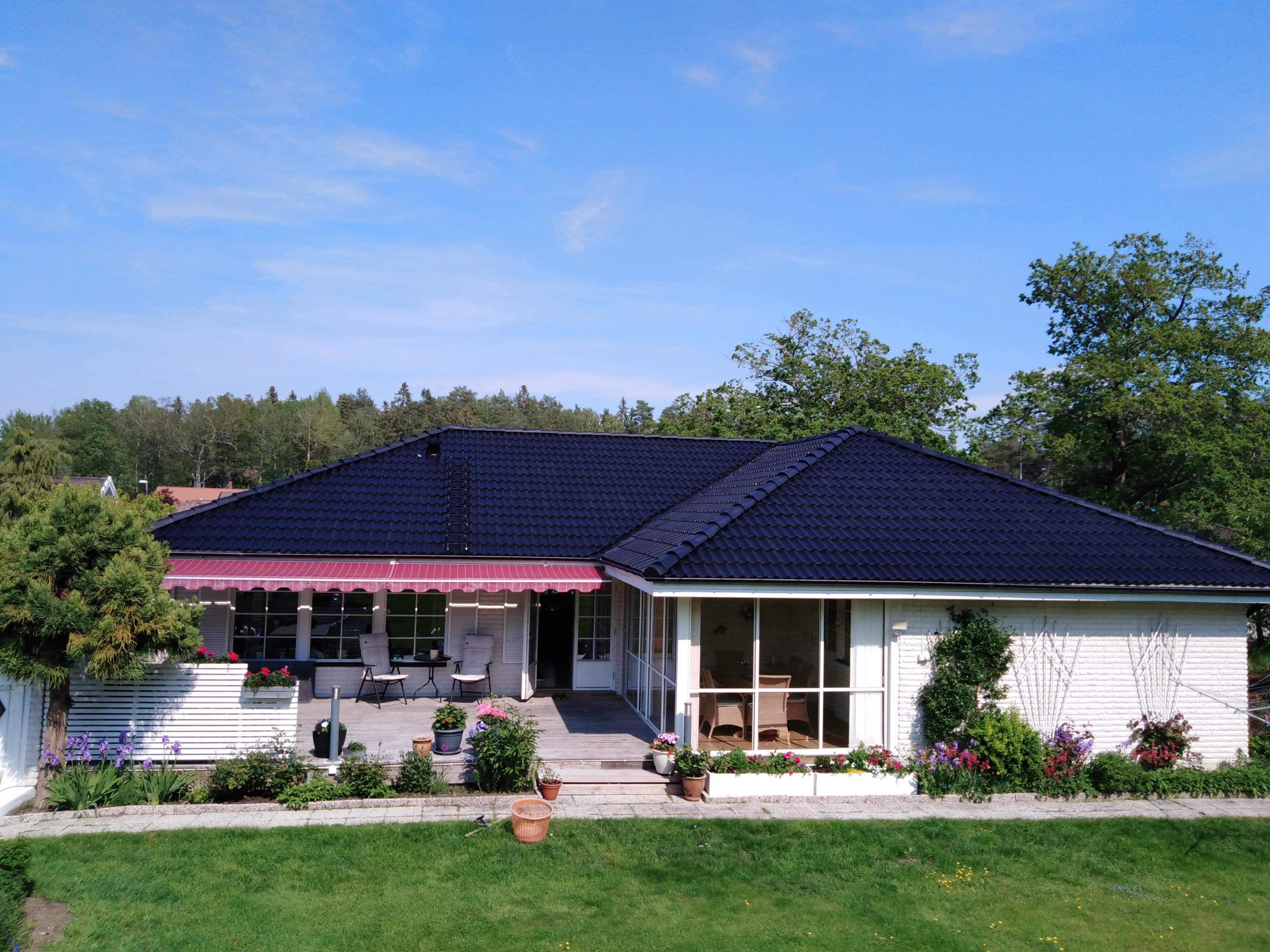 Widok z lotu ptaka na dom z ciemnym dachem, białymi ścianami i czerwono-białą markizą nad tarasem, otoczony zielenią trawnika, drzew i kwiatów.