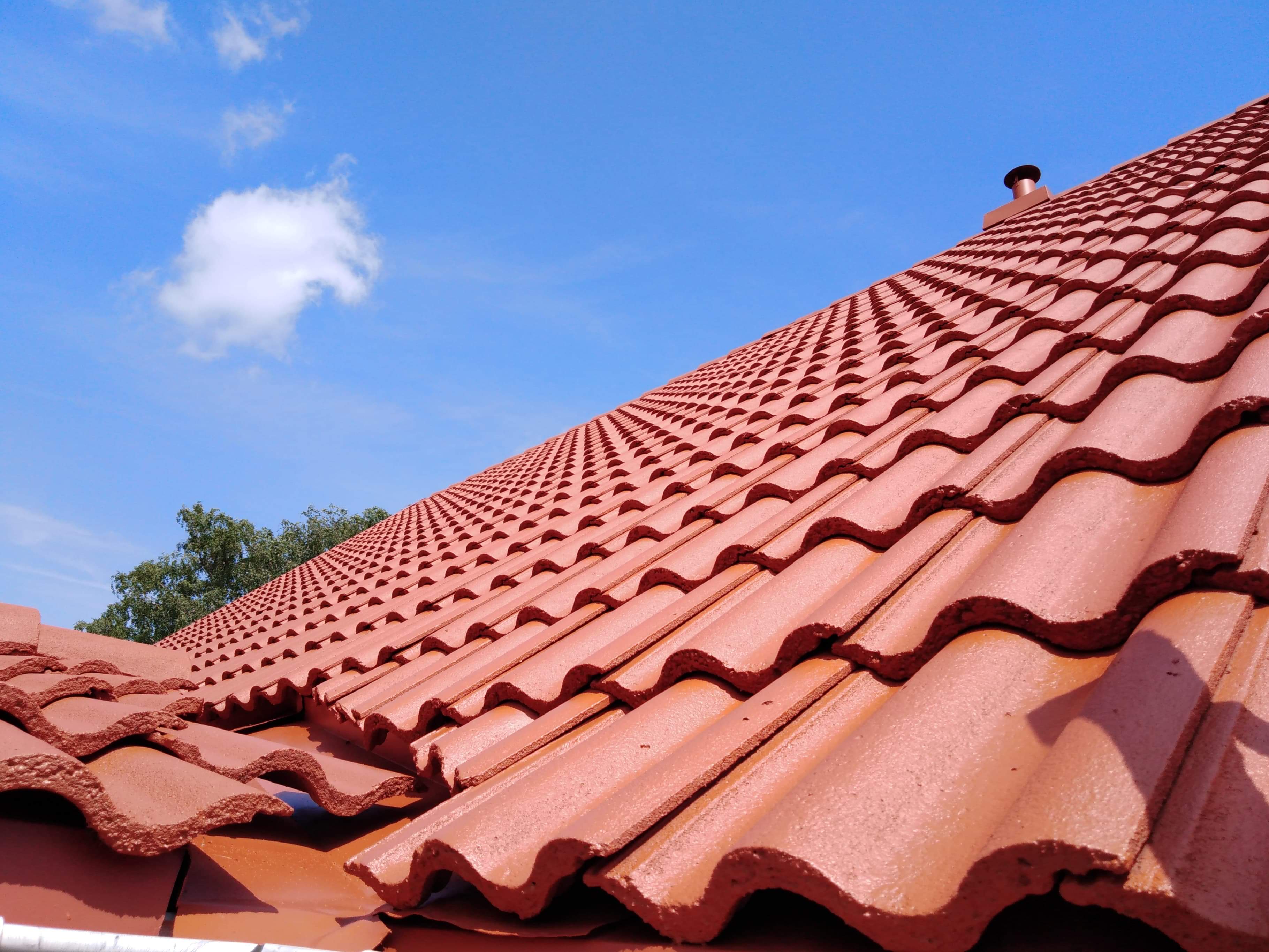 Nowo pomalowany dach z czerwonej dachówki ceramicznej, widok z dołu na tle błękitnego nieba z chmurą, widoczny komin wentylacyjny.