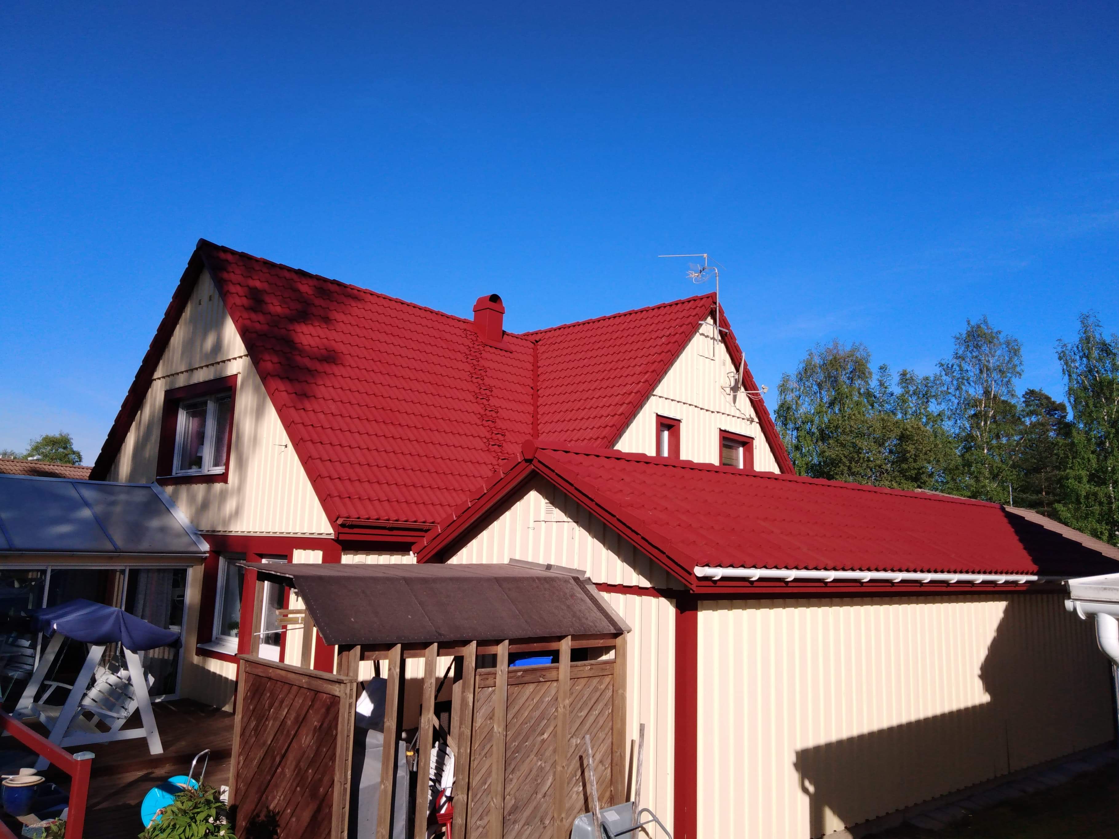 Dom z nowym, czerwonym dachem z blachy falistej, kremowa elewacja z czerwonymi akcentami, antena na dachu, błękitne niebo w tle.