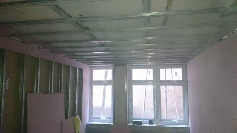 Montaż metalowego rusztu pod płytę gipsowo-kartonową na suficie i ścianach w pomieszczeniu z dwoma oknami.