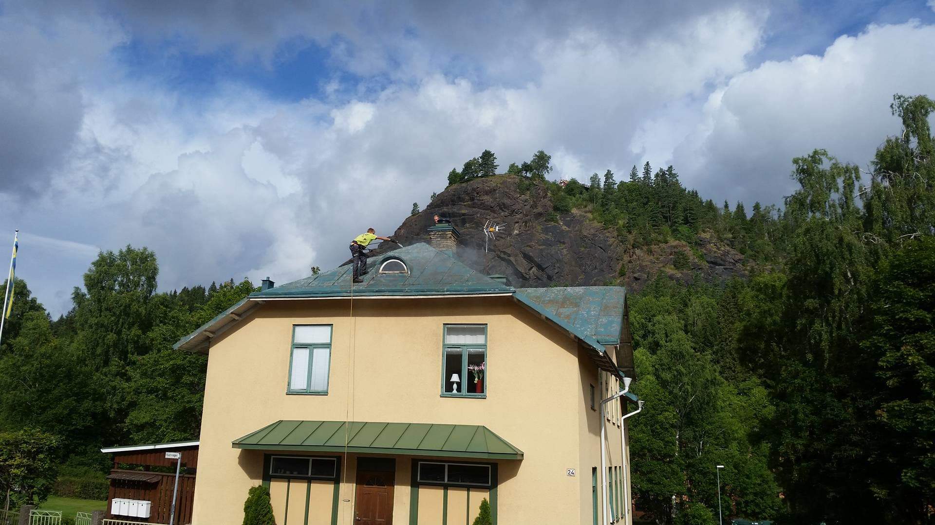 Pracownik w żółtej koszulce czyści dach budynku myjką ciśnieniową, z widocznym wzgórzem w tle.