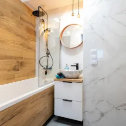 Nowoczesna łazienka z drewnopodobnymi i marmurowymi płytkami, okrągłym lustrem i czarną armaturą.