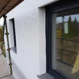 Fragment elewacji budynku z oknami, pokrytej jasnym, ziarnistym tynkiem, z widocznymi fragmentami rusztowania.