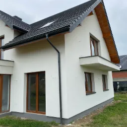 Nowoczesny dom jednorodzinny z białą elewacją, ciemną dachówką i drewnianymi elementami wykończeniowymi, widok z boku w pochmurny dzień.