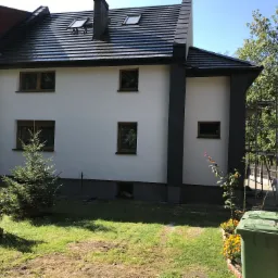 Dwukondygnacyjny dom z białą elewacją i ciemnoszarymi akcentami, widoczne okna z brązowymi ramami, czarny dach z oknami dachowymi, zielony trawnik z krzewami i drzewami, metalowe rusztowanie z boku...