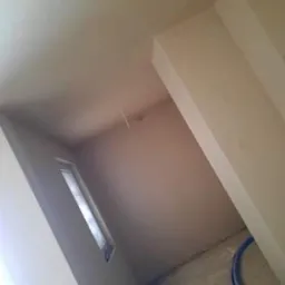 Świeżo położone tynki maszynowe na ścianach i suficie, widoczna listwa przyścienna i okno z widokiem na zewnątrz, przewód instalacyjny przy podłodze.