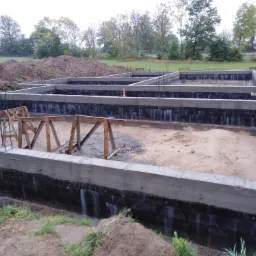 Fundamenty domu jednorodzinnego z hydroizolacją, widoczne drewniane podpory szalunku i wystające pręty zbrojeniowe, w tle hałdy ziemi i drzewa.