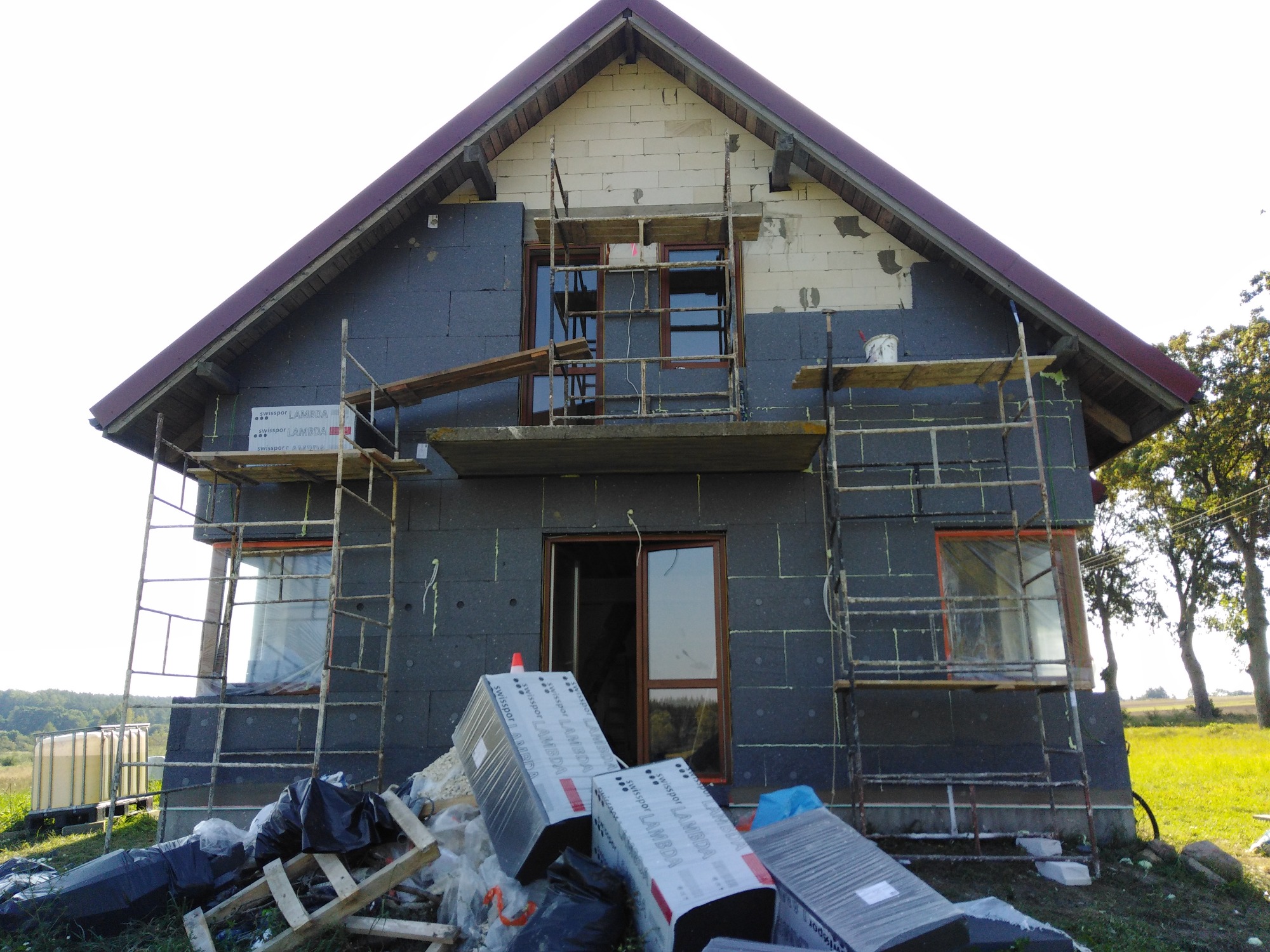 Dom w trakcie ocieplania styropianem Swisspor Lambda, widoczne rusztowania, częściowo ocieplona ściana szczytowa, nieotynkowane ściany z pustaków, paczki styropianu i worki z odpadami budowlanymi...