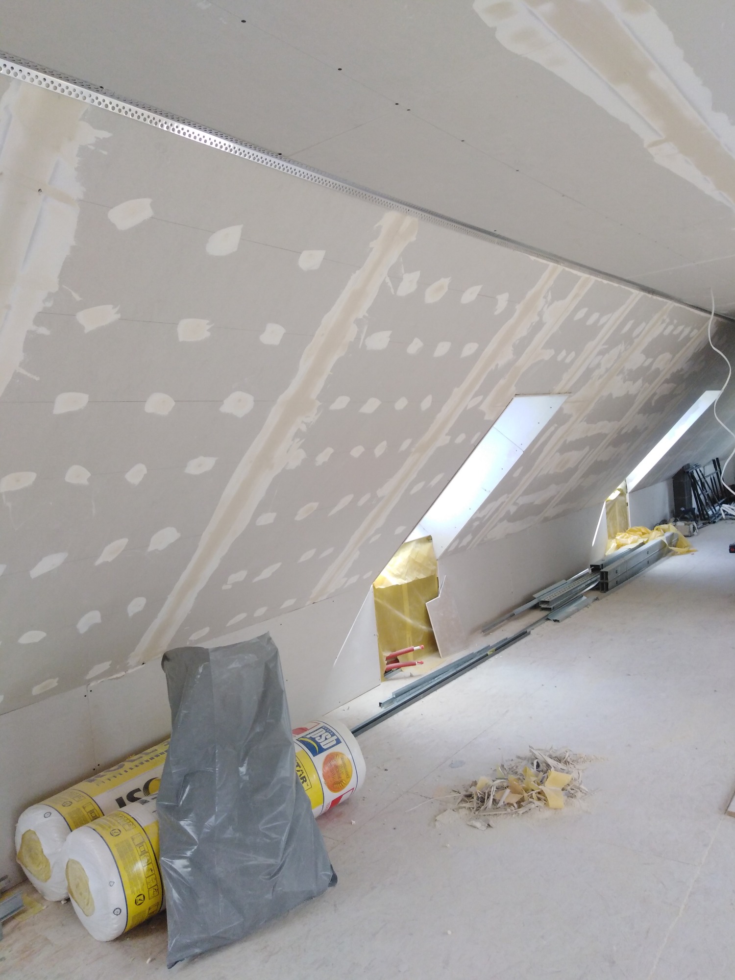 Skośny sufit poddasza w trakcie szpachlowania, widoczne punkty po wkrętach, łączenia płyt gipsowo-kartonowych, profile metalowe, okna dachowe, rolki wełny mineralnej i worek na ścinki.