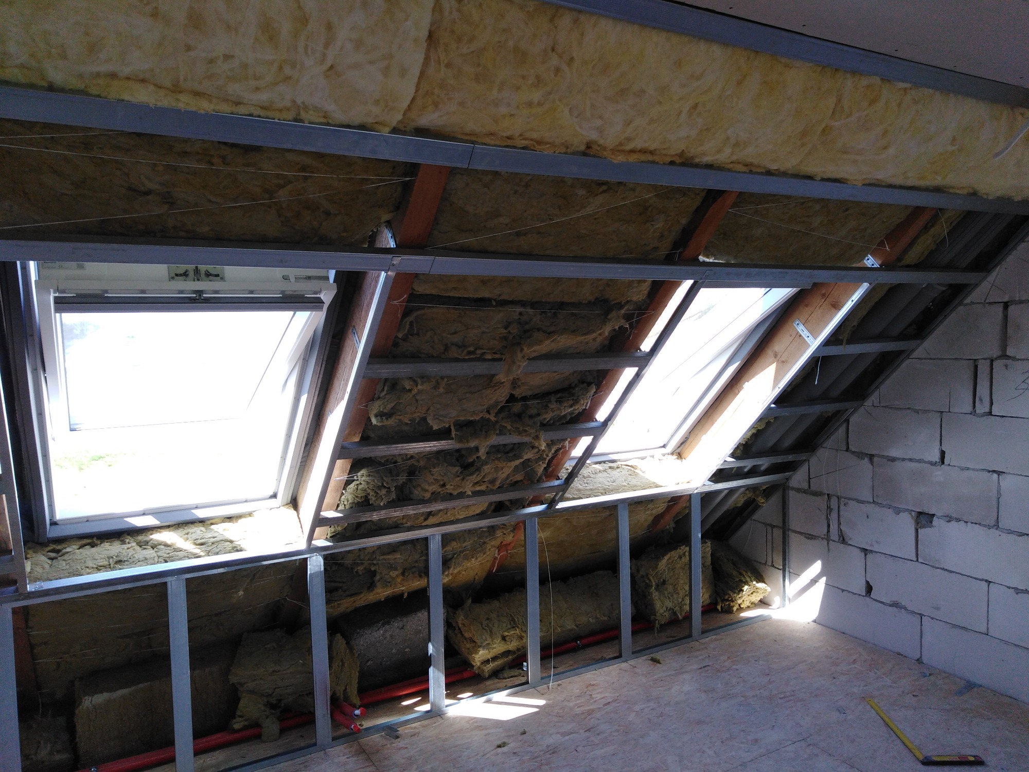 Poddasze w trakcie prac izolacyjnych: widoczne okna dachowe, wełna mineralna w metalowej konstrukcji, surowy mur z bloczków betonowych, fragment podłogi i instalacji grzewczej.