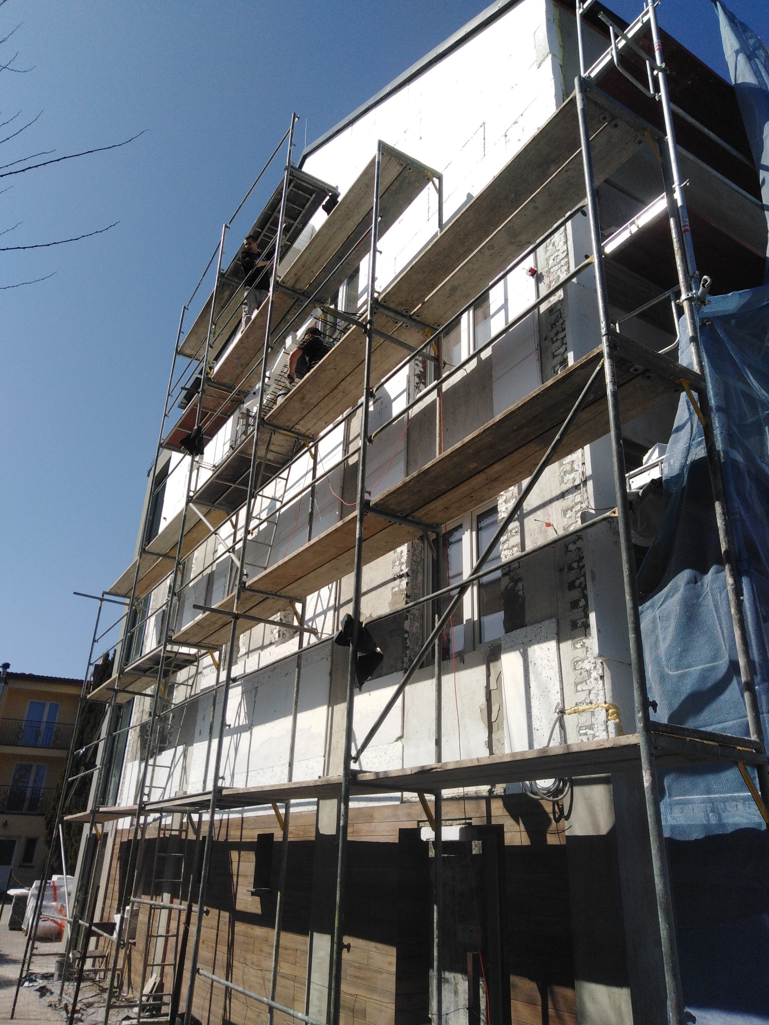 Budynek w trakcie ocieplania, widoczne rusztowania, pracownicy na wysokościach, ściany częściowo pokryte izolacją, fragmenty elewacji odsłonięte.