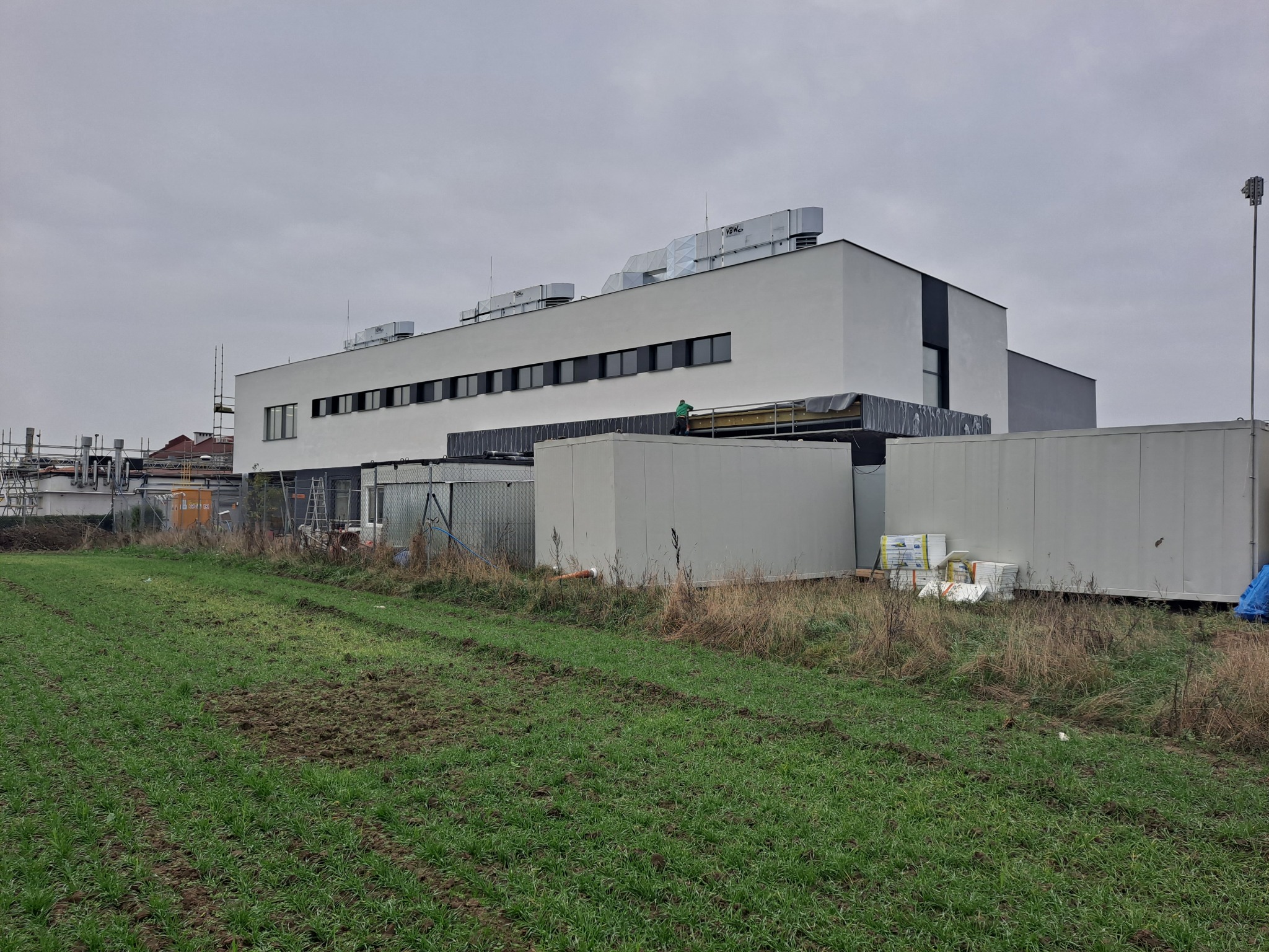 Nowoczesny, dwukondygnacyjny budynek biurowy z białymi ścianami i czarnymi ramami okiennymi, widoczny z perspektywy pola, z kontenerami budowlanymi na pierwszym planie i osobą pracującą na dachu.