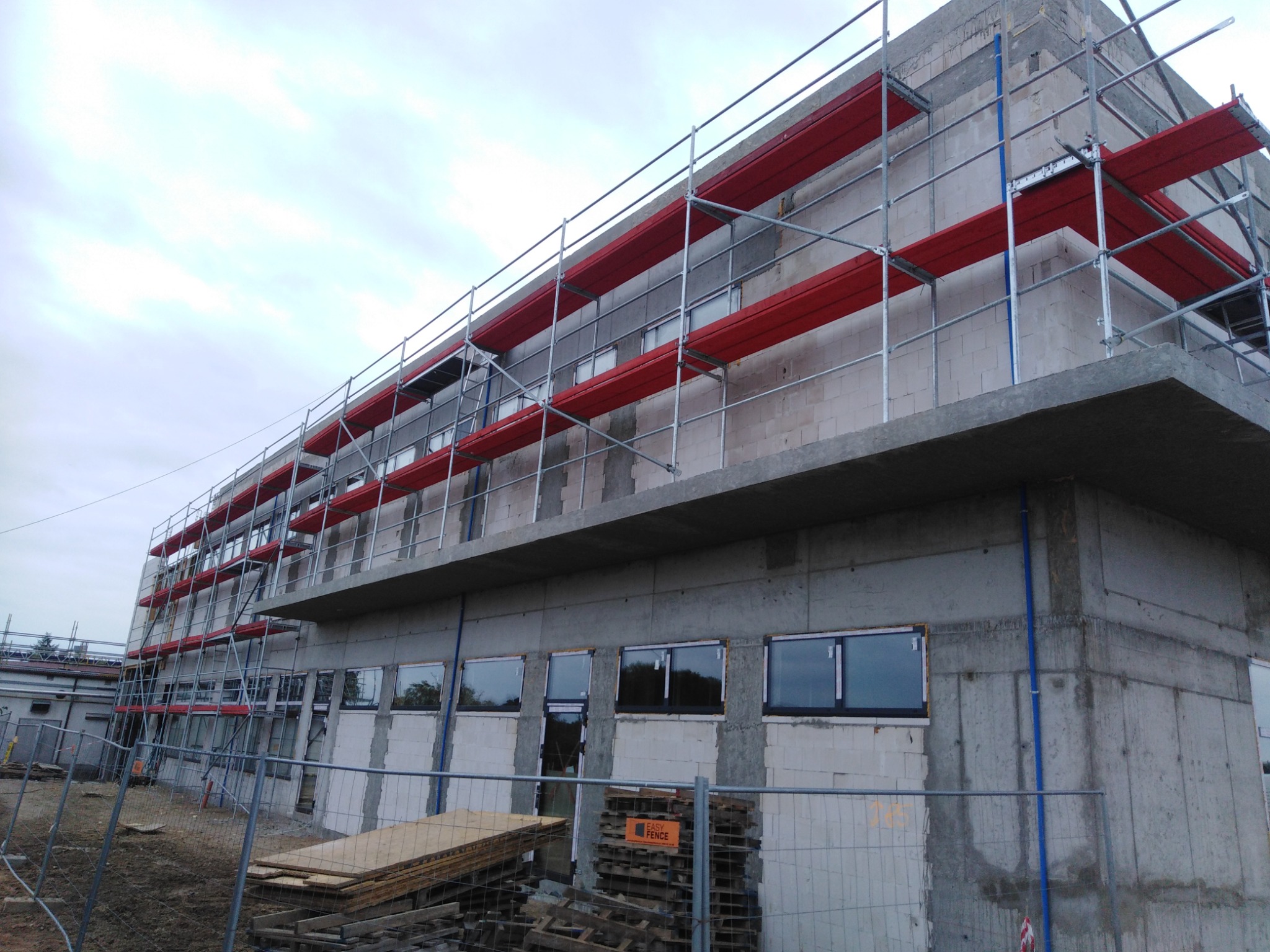 Budynek wielopiętrowy w trakcie prac elewacyjnych, z widocznymi rusztowaniami z czerwonymi podestami i surowymi ścianami z betonu oraz bloczków, zabezpieczony siatką ogrodzeniową.
