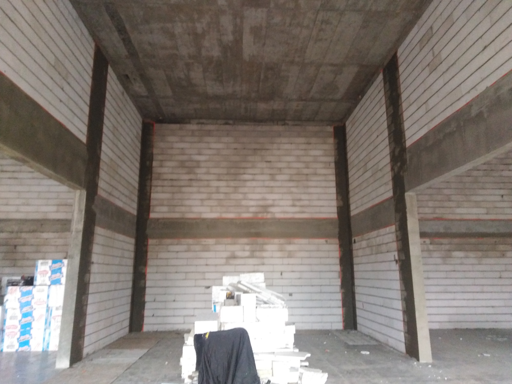 Wnętrze w trakcie prac budowlanych: ściany z białych bloczków, surowy betonowy strop, widoczne wzmocnienia konstrukcyjne, materiały budowlane składowane wewnątrz.