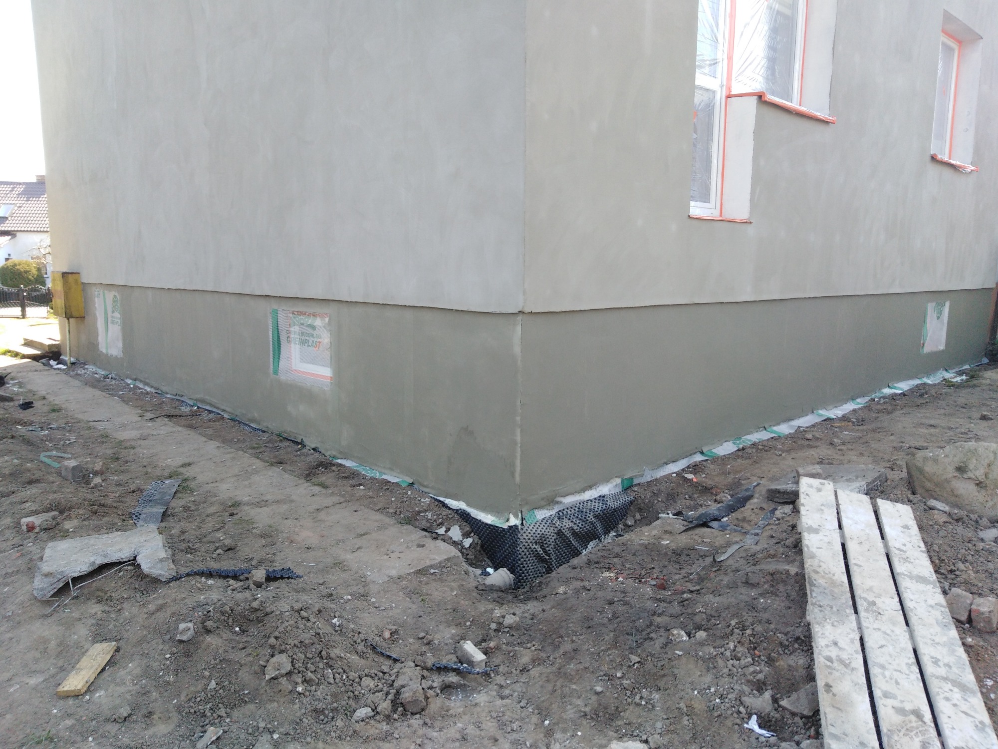 Fragment ocieplonej elewacji domu jednorodzinnego z widocznym fragmentem fundamentu, zabezpieczonym folią kubełkową oraz oknami zabezpieczonymi folią malarską.