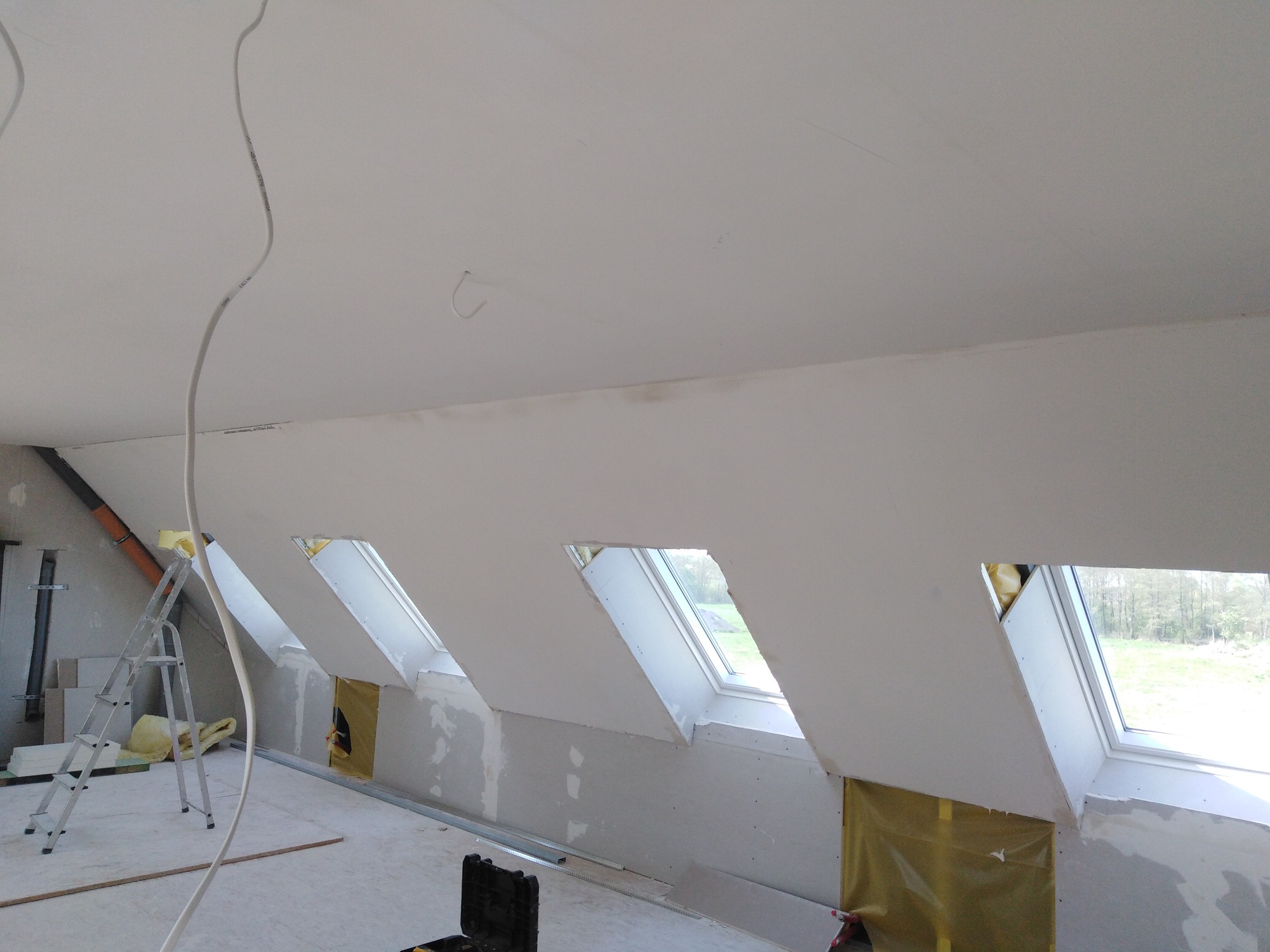 Poddasze w Kędrzynie w trakcie prac wykończeniowych: zamontowane płyty gipsowo-kartonowe na skosach z oknami dachowymi, widoczna drabina i materiały izolacyjne.