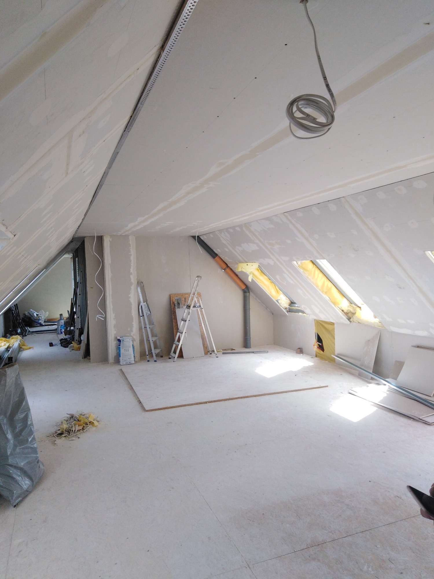 Poddasze w trakcie wykańczania, widoczne płyty karton-gips na skosach dachu i ścianach, przygotowane podłoże pod dalsze prace, w oknach dachowych widoczna izolacja.