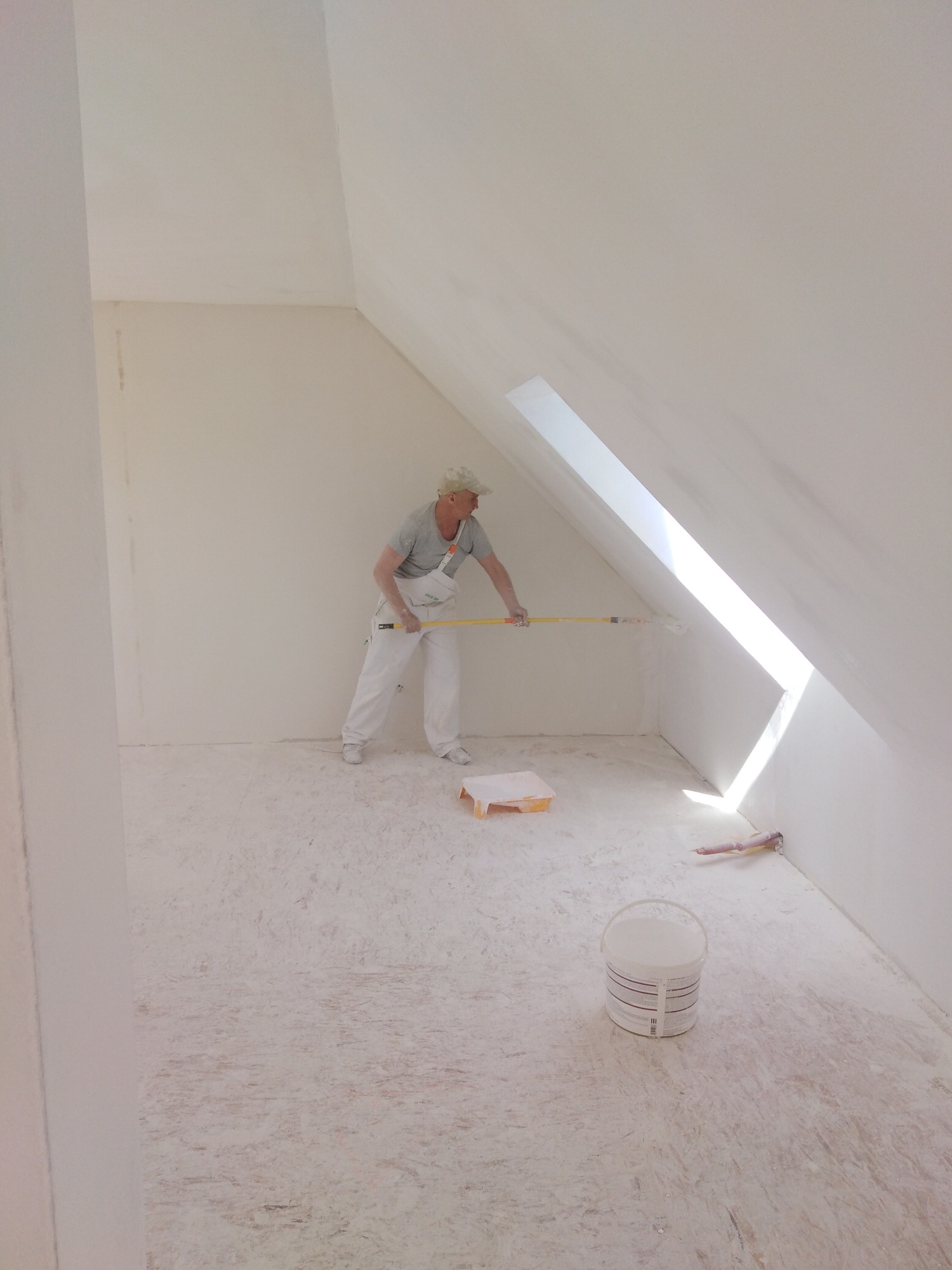 Pracownik w kombinezonie maluje ścianę w pomieszczeniu ze skosami, używając wałka na długim kiju. Na podłodze widoczne ślady farby, wiadro farby i kuweta.