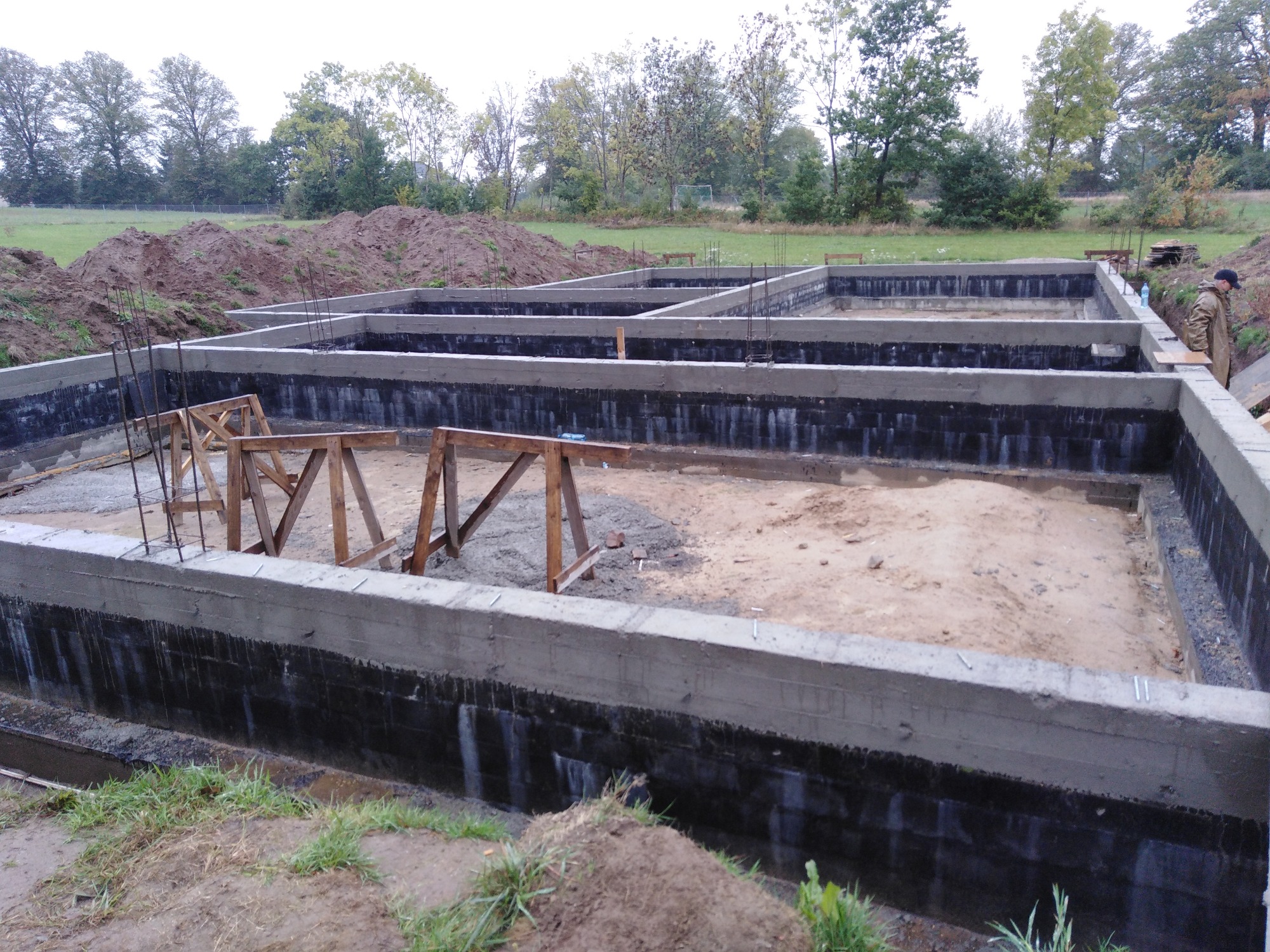 Fundamenty domu jednorodzinnego z hydroizolacją, widoczne drewniane podpory szalunku i wystające pręty zbrojeniowe, w tle hałdy ziemi i drzewa.