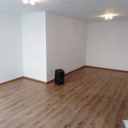 Puste, jasne pomieszczenie po remoncie z drewnopodobnymi panelami podłogowymi, białymi ścianami i widocznym grzejnikiem elektrycznym przy ścianie.