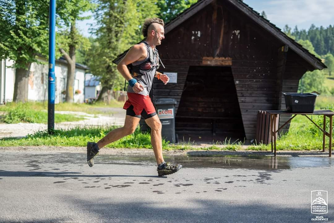 Mężczyzna z irokezem biegnie w zawodach górskich, ubrany w sportową koszulkę, czerwone spodenki i buty trailowe, obok drewnianej wiaty i kałuż na asfalcie, widoczny logotyp imprezy sportowej.