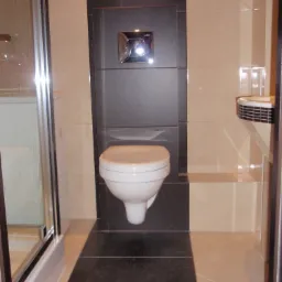 Nowoczesna toaleta podwieszana z chromowanym przyciskiem spłukującym, obudowana czarnymi płytkami, w łazience wykończonej beżowymi płytkami i kabiną prysznicową z przezroczystego szkła.