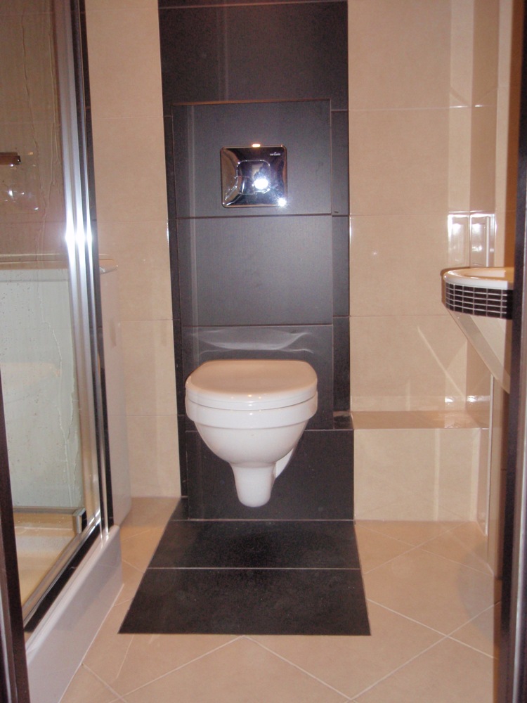 Nowoczesna toaleta podwieszana z chromowanym przyciskiem spłukującym, obudowana czarnymi płytkami, w łazience wykończonej beżowymi płytkami i kabiną prysznicową z przezroczystego szkła.