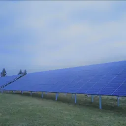 Dwa rzędy paneli słonecznych na metalowych stelażach, ustawione na trawiastym terenie; w tle budynki mieszkalne pod pochmurnym niebem.