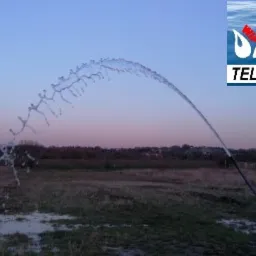 Test wydajności studni w Zborowicach: strumień wody w kształcie łuku na tle zachodzącego słońca i łąki, widoczne logo firmy z numerem telefonu.