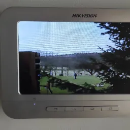 Zamontowany na ścianie panel wideodomofonu Hikvision wyświetlający obraz z kamery monitoringu na zewnątrz, widoczny ogród z osobą.