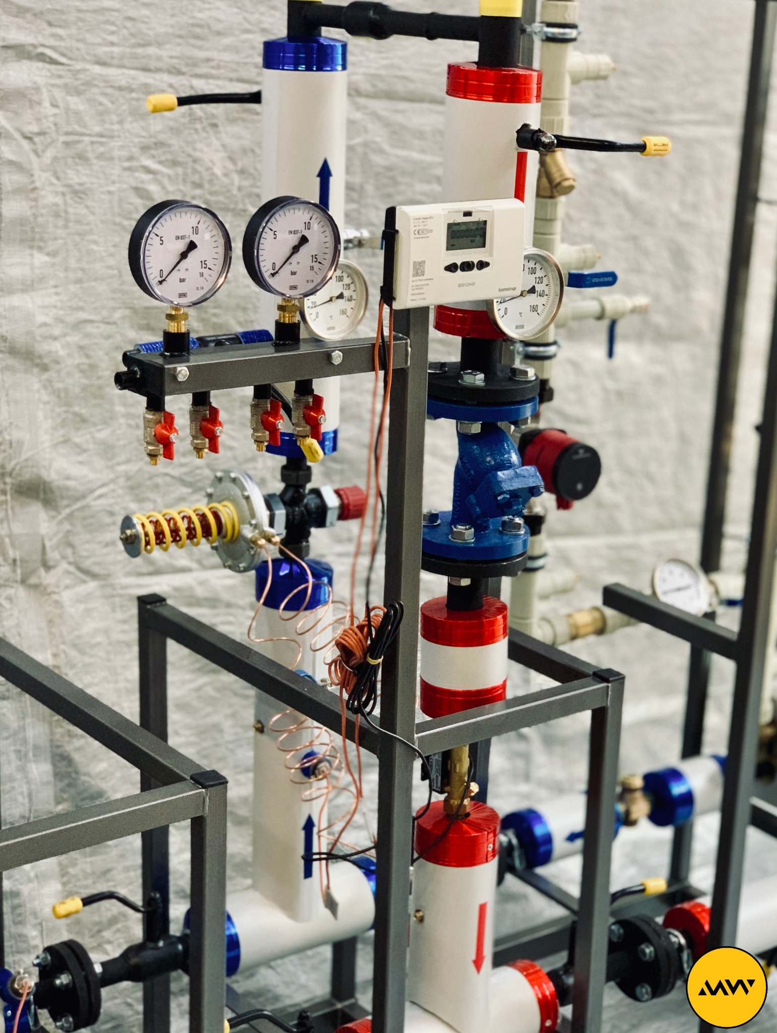 Rozbudowana instalacja grzewcza z manometrami, zaworami i licznikiem ciepła, zamontowana na stalowej ramie. Widoczne rury z oznaczeniami kierunku przepływu, kolorowe elementy (czerwone, niebieskie,...