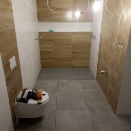 K.M.D. Mariusz Ratajczak - Wnętrze łazienki w trakcie remontu: ułożone płytki imitujące drewno na ścianach, szare płytki podłogowe, wisząca toaleta z narzędziami na górze, kable zwisające z sufitu.