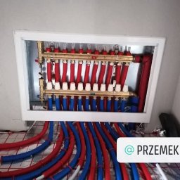 Mariusz Ratajczak Zaklad instalacji sanitarnych - Rozdzielacz ogrzewania podłogowego z czerwono-niebieskimi rurami, zamontowany w białej skrzynce, widoczne elementy instalacji na podłodze.