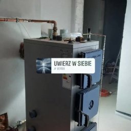 K.M.D. Mariusz Ratajczak - Szary piec CO z niebieskimi drzwiczkami, miedziane rury instalacji grzewczej na ścianie, surowy stan wykończenia wnętrza. Widoczne akcesoria na piecu.