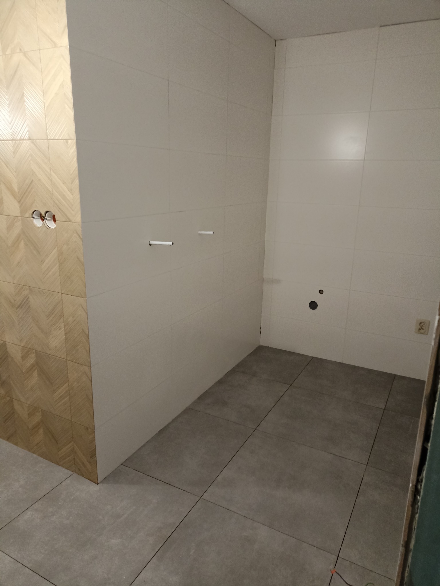 Wykończona łazienka z szarymi płytkami podłogowymi i białymi ściennymi. Widoczne przygotowane punkty hydrauliczne i elektryczne. Część ściany wykończona płytkami imitującymi drewno.