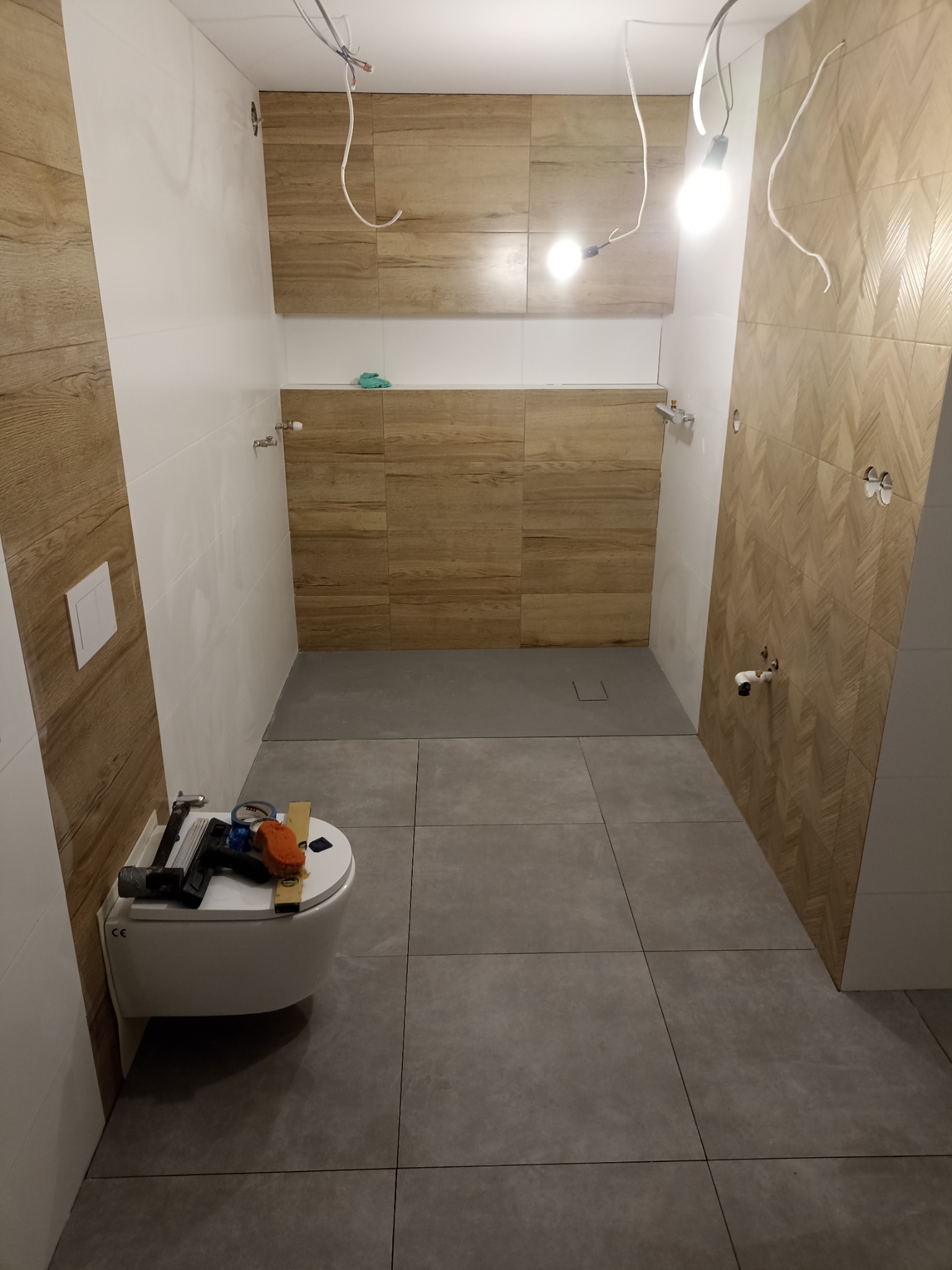 Wnętrze łazienki w trakcie remontu: ułożone płytki imitujące drewno na ścianach, szare płytki podłogowe, wisząca toaleta z narzędziami na górze, kable zwisające z sufitu.