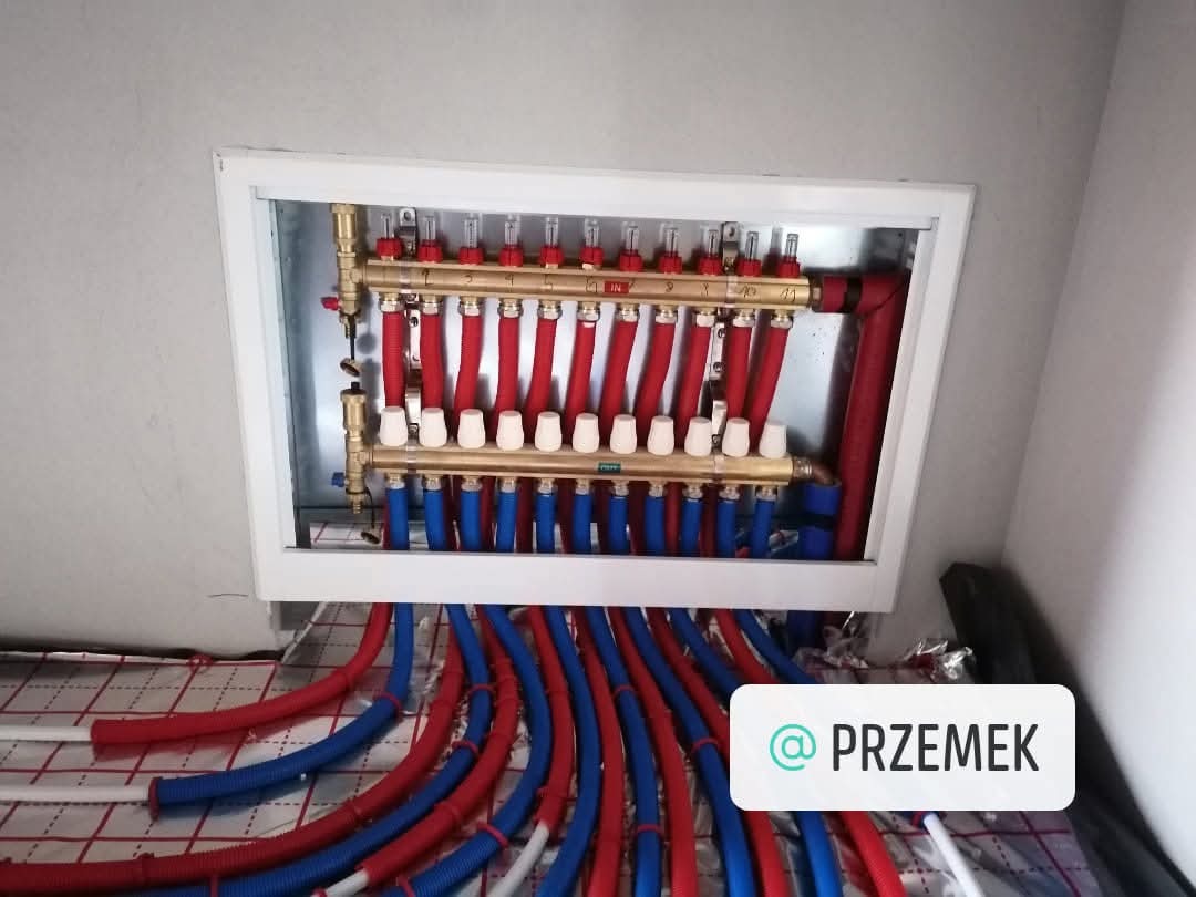 Rozdzielacz ogrzewania podłogowego z czerwono-niebieskimi rurami, zamontowany w białej skrzynce, widoczne elementy instalacji na podłodze.