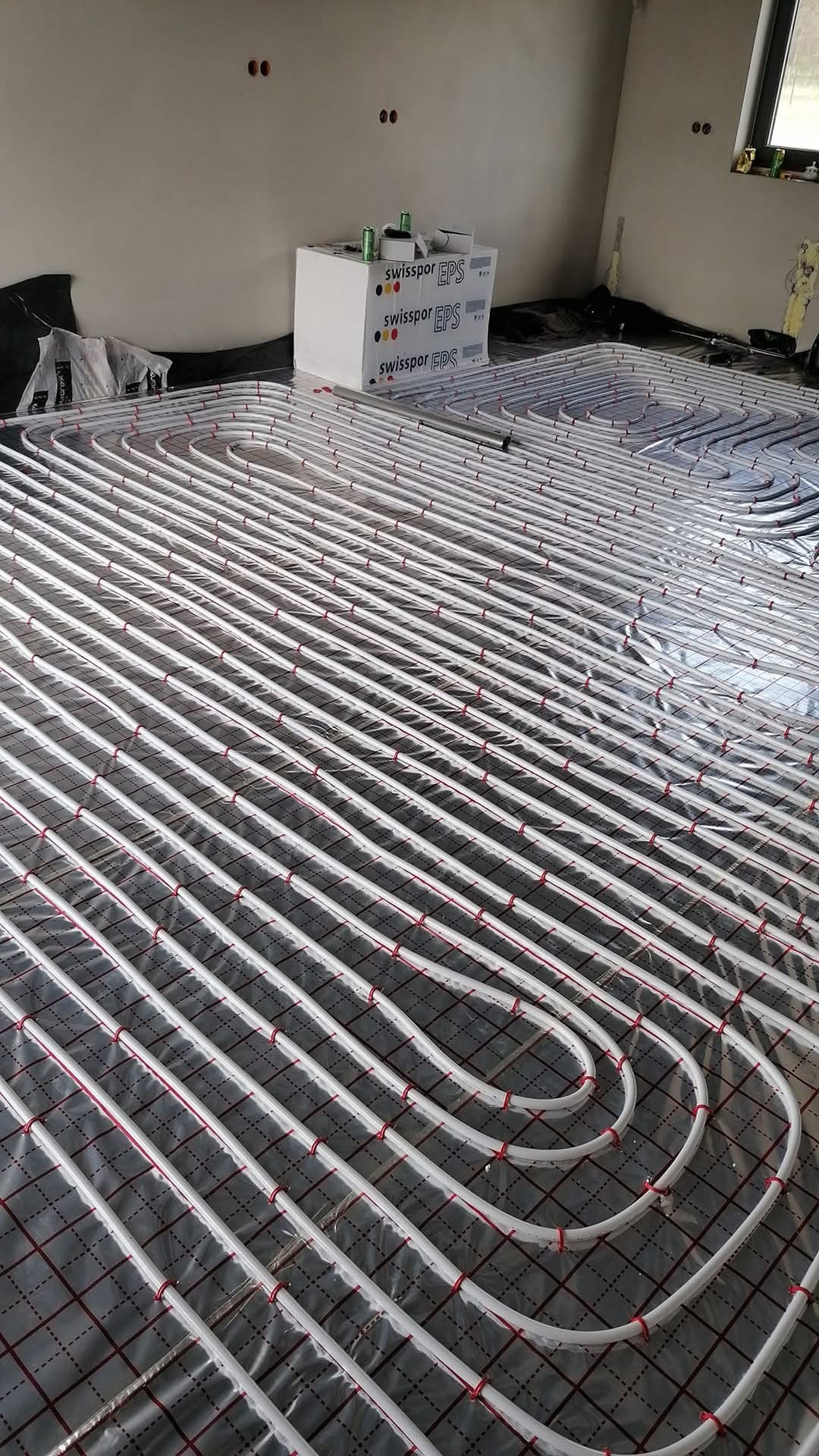 Białe rury ogrzewania podłogowego ułożone na folii z czerwoną siatką, widoczne puszki i styropian. Instalacja w surowym wnętrzu przed wylaniem posadzki.
