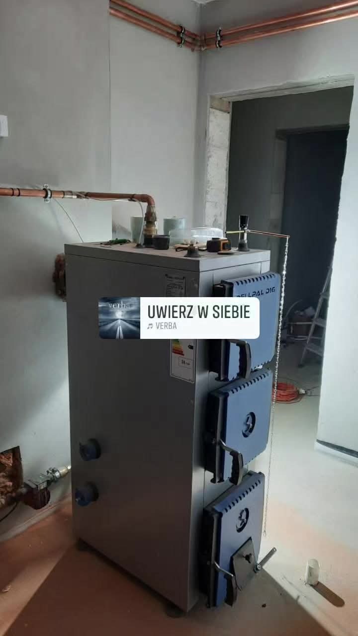 Szary piec CO z niebieskimi drzwiczkami, miedziane rury instalacji grzewczej na ścianie, surowy stan wykończenia wnętrza. Widoczne akcesoria na piecu.