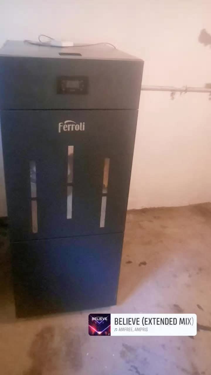 Stojący, szary piec CO marki Ferroli z widocznym panelem sterowania i otworami wentylacyjnymi, umieszczony w pomieszczeniu z surowym wykończeniem podłogi.