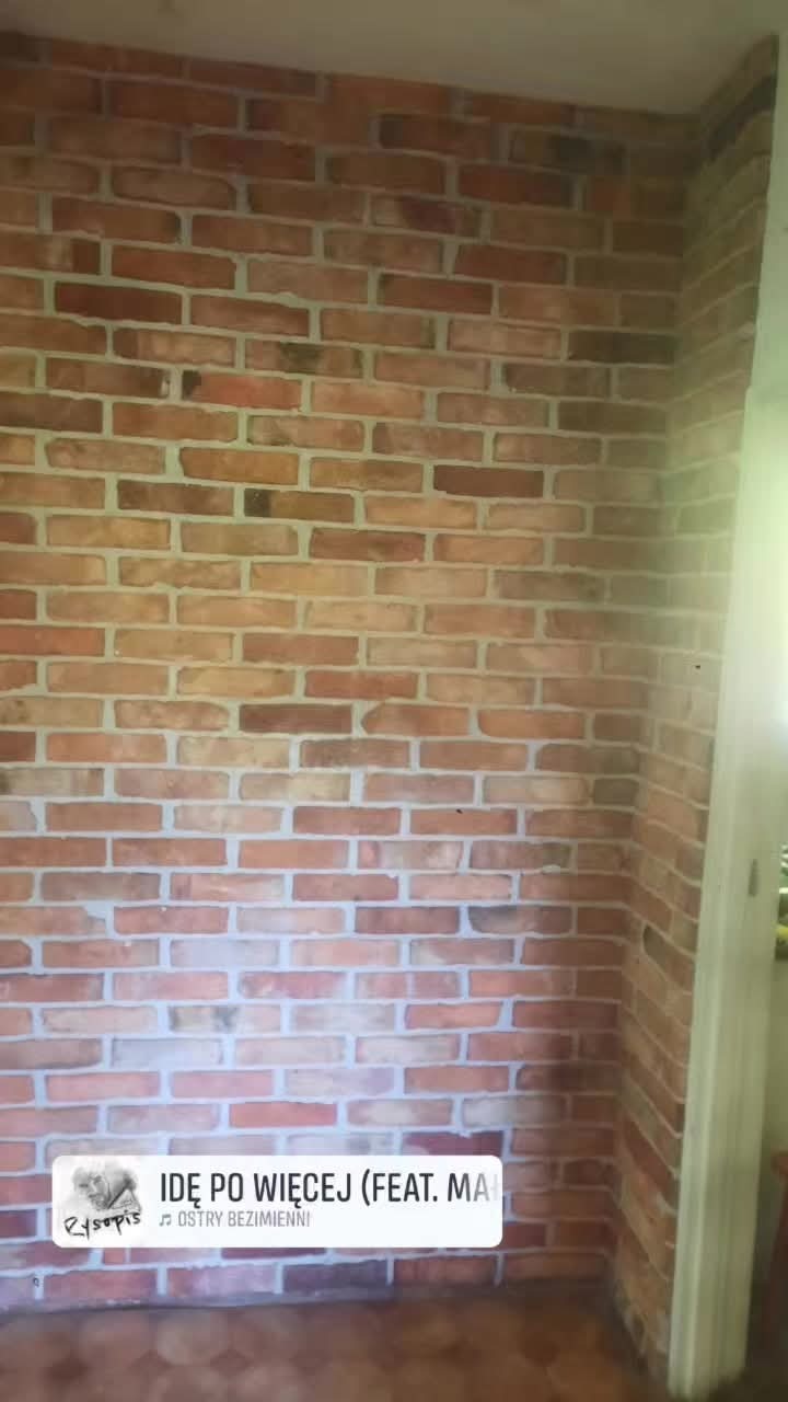 Ściana z cegły w odcieniach czerwieni i brązu, z widoczną fugą. Fragment ściany z widocznym oświetleniem i fragmentem podłogi. Dekoracyjna ściana z cegły.