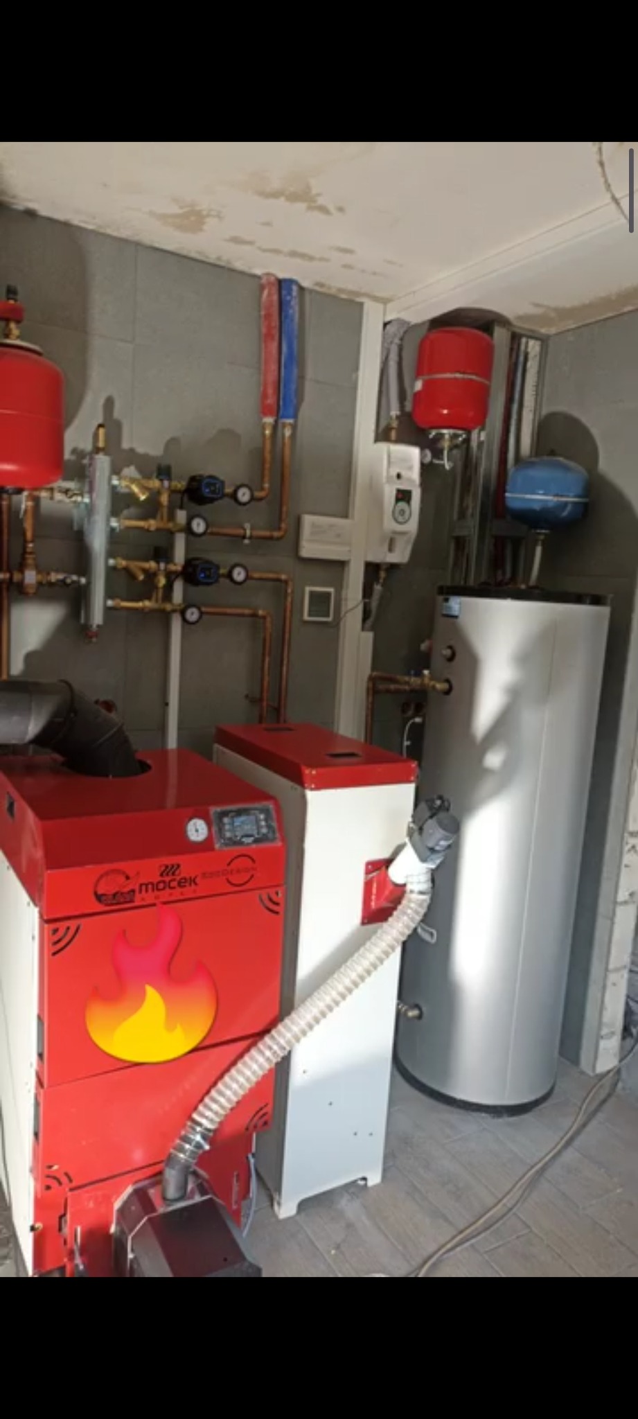 Kotłownia z piecem na paliwo stałe, zasobnikiem ciepłej wody i instalacją CO w pomieszczeniu wykończonym płytkami. Widoczne rury miedziane i elementy sterujące.