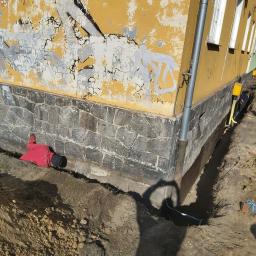 DAR-KOP - Pracownik w czerwonej kurtce w wykopie przy fundamencie budynku z żółtą elewacją i kamienną podmurówką, widoczne rury instalacyjne i skrzynki przyłączeniowe.